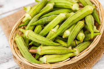 Wooden bowl filled with fresh okra pods placed on a piece of burlap fabric on a rustic wooden white surface.