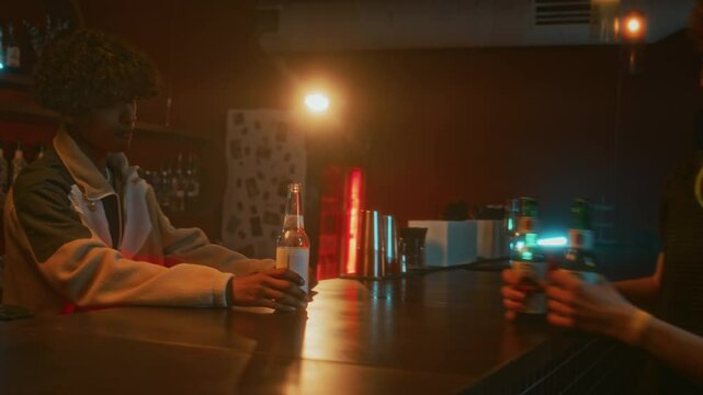 Side shot of cool raver guy in trendy outfit and techno sunglasses returning empty bottle of beer to young bartender and taking two more while hanging out in nightclub