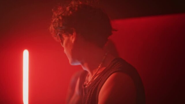 Chest up handheld shot of confident charismatic guy in techno rave sunglasses and y2k outfit moving energetically to electronic dance music in smoky red neon lit nightclub