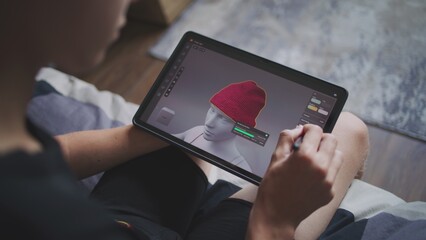Boy with digital stylus and tablet in hands sits at home, creates 3d models. Focused teenager draws clothes for game character using 3d modeling program. Development and game design concept. Close up.