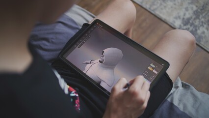 Focused teenager with digital pencil and tablet in hands sits in room, draws 3d model. Boy creates new game character and animation using 3d modeling program. Game design and development. Close up.