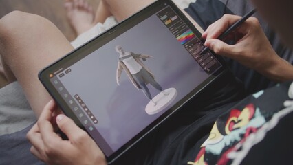 Focused teenager draws clothes for game character using 3d modeling program. Boy with digital pencil and tablet in hands sits in room, creates 3d models. Game design and development concept. Close up.