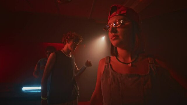 Handheld lowkey shot of young confident lady in denim top, cap and oval sunglasses making relaxed movements to electronic dance music during retro party in red neon lit nightclub