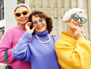 Three trendy elderly women wearing colorful sweaters and sunglasses posing for the camera in front of an office building,