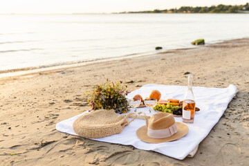 Cozy beach picnic on a white blanket with fresh summer fruits, lemonade, wine, straw hat on the beach by the sea and ocean
