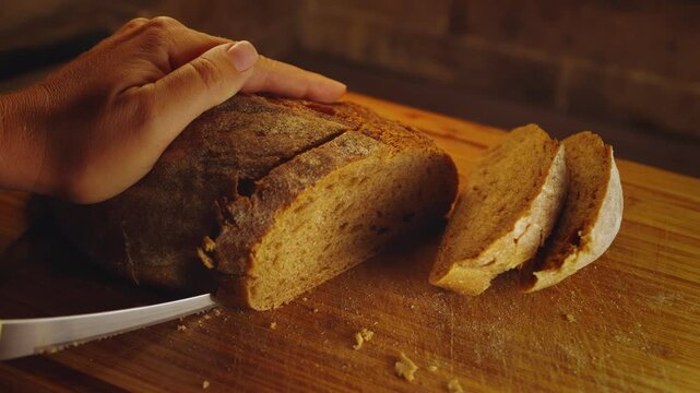 Close-up hands of female baker with knife cutting freshly baked aromatic loaf of wholemeal bread to slices at bakery or kitchen on chopping board