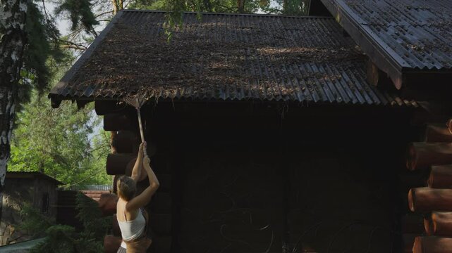 Female worker carries out maintenance of a private home clears the roof of a house from leaves and branches