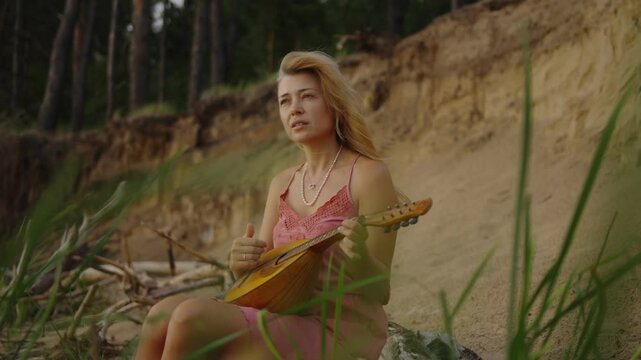 Attractive young woman sitting in nature on the sandy shore playing mandolin