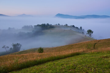 Beautiful dreamy autumn sunrise rural scenery. Haystacks and trees on a mountain hill with fog.