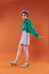 A stylish woman in a green blouse and lavender skirt poses playfully in a vibrant orange studio.