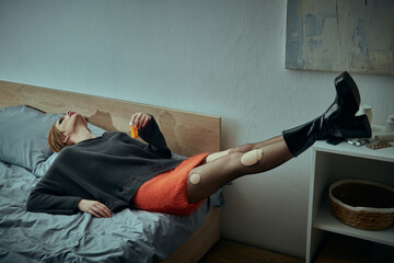 A young woman in a chic grungy style lies on a bed, legs raised, with a relaxed expression.