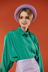 A stylish woman poses in a green shirt and lavender hat, against a warm orange background.