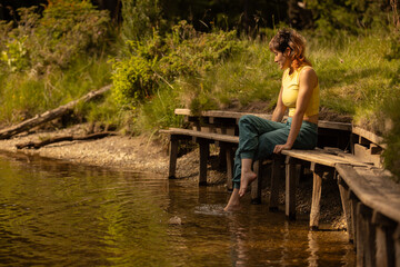 Fototapeta premium A woman sits on a bench by a body of water, her feet in the water
