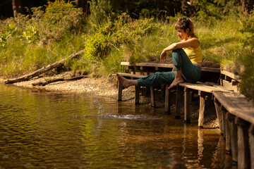 A woman sits on a wooden pier by a lake, with her feet in the water