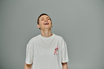 A woman with short hair, wearing casual clothing and a pink ribbon, smiles brightly against a grey backdrop.