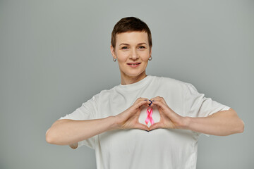 Woman with short hair, white shirt and pink ribbon forms a heart with hands, showing support for...