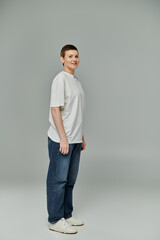 Short-haired woman in jeans, white T-shirt, and sneakers smiles confidently against a grey backdrop in casual attire