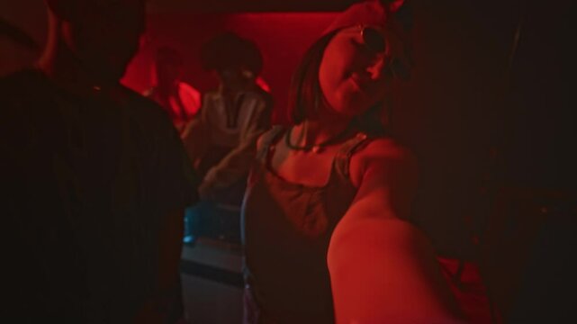 POV handheld lowkey shot of young cool lady recording herself on front camera while dancing with stylish guy during retro party in nightclub with red neon light and flashes
