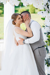 A bride and groom are kissing each other in front of a white arch. The bride is wearing a white dress and the groom is wearing a gray vest. Scene is romantic and intimate