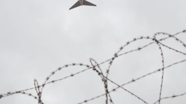 Looking up view white passenger airplane in cloudy sky, airport perimeter fence foreground. Focus on aircraft overhead bottom view, barbwire bokeh blurred. Barded wire, flying commercial plane