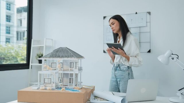 Beautiful caucasian architect focus on using tablet to check house model on meeting table with blueprint, house model and architectural document scatter around at meeting room. Immaculate.