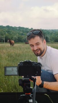 Vertical footage of a young vlogger films himself against a background of horses in a field and says something with a smile.