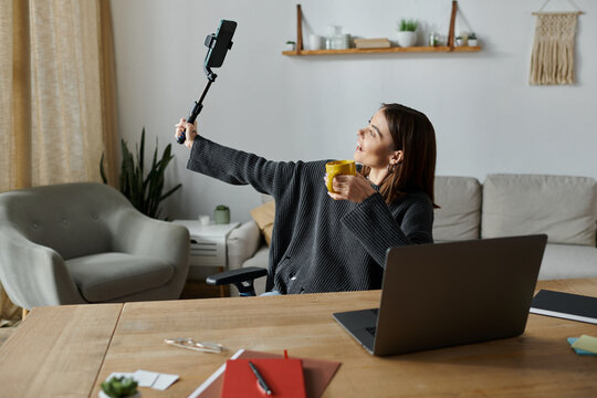 A young woman in a grey sweater films content with a selfie stick while sipping from a yellow mug. - Powered by Adobe