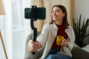 A young woman in casual attire sits on a couch, smiling as she vlogs while holding a smartphone stabilizer.