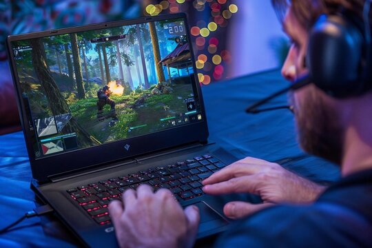 Close-up of a man intensely playing a shooter video game on his laptop at home in a forest-themed virtual environment.