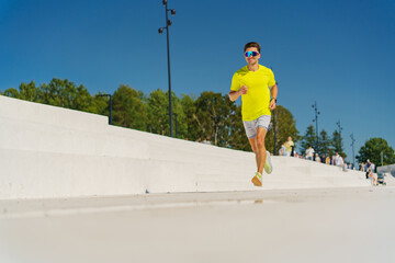 Focused jogger in bright attire running along sunlit steps, surrounded by trees and clear skies.