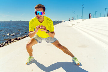 Smiling athlete in bright sportswear performs a stretch by the waterfront under a clear blue sky.