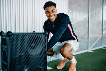 Smiling young man in sportswear turning on a large wireless speaker to play some music before a workout session at the gym