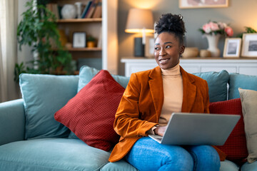 Beautiful African woman sitting on a couch at home and using laptop computer, smiling and looking aside.