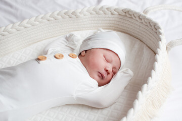 A newborn baby girl sleeping in a Moses basket in a white sleeping sack and a white hat
