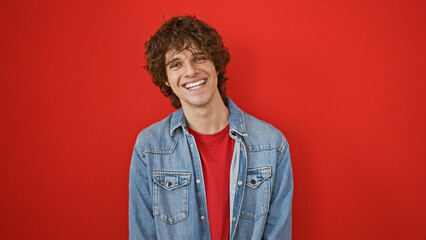 Smiling young hispanic man with curly hair, beard, and denim jacket against a vibrant red wall background.