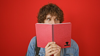 Curly-haired man peers over red notebook against a vibrant red background, conveying curiosity and study.