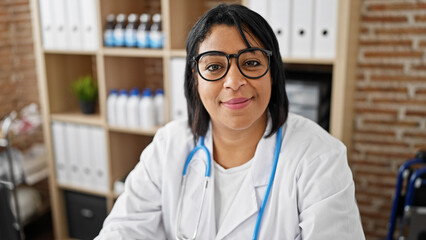 Hispanic woman wearing glasses and lab coat with a stethoscope in a medical clinic office smiling at the camera