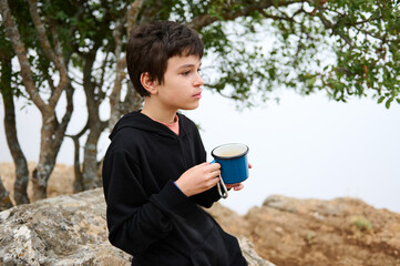 Young boy enjoying a hot drink in nature, wearing a black hoodie seated on a rock