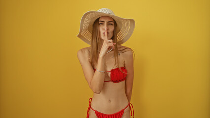 Beautiful blonde woman in a red bikini and straw hat making a shushing gesture, isolated over a...