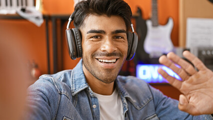 Smiling young hispanic man with beard wearing headphones in a music studio waving at the camera for...