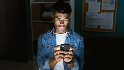 A young hispanic man in a denim jacket using a smartphone indoors with an office background.
