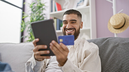 Fototapeta premium Smiling man with beard holding tablet and credit card indoors on sofa for online shopping or banking.