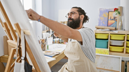 A bearded artist painting on a canvas in a bright studio with art supplies visible