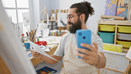 Young bearded man in apron taking selfie in art studio with paintings and supplies around him.