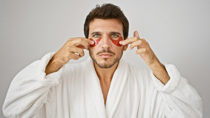 Handsome bearded young man in a white robe applying red under-eye patches against an isolated white background.