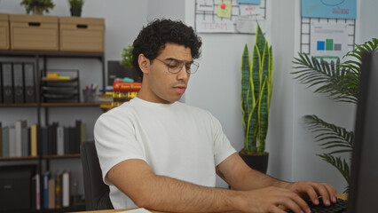 Hispanic man working in an office, focused on his computer, surrounded by organized shelves and green plants in a modern workplace.