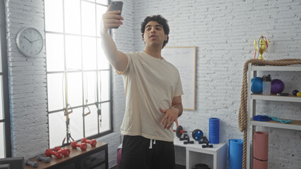 Young hispanic man taking selfie in a modern gym with equipment around him