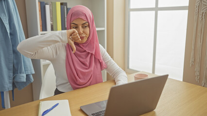 Young hispanic woman in hijab making thumbs-down gesture at home office with laptop and notebook.