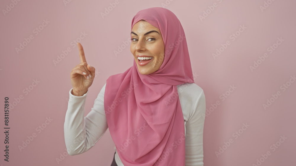 Wall mural A smiling young woman wearing a hijab points upwards, standing against a soft pink background, embodying positivity and beauty.