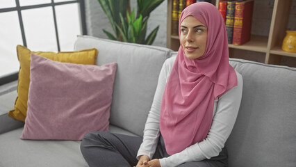 Fototapeta premium A thoughtful woman in a pink hijab rests on a grey sofa in a cozy living room, evoking a sense of contemplation.